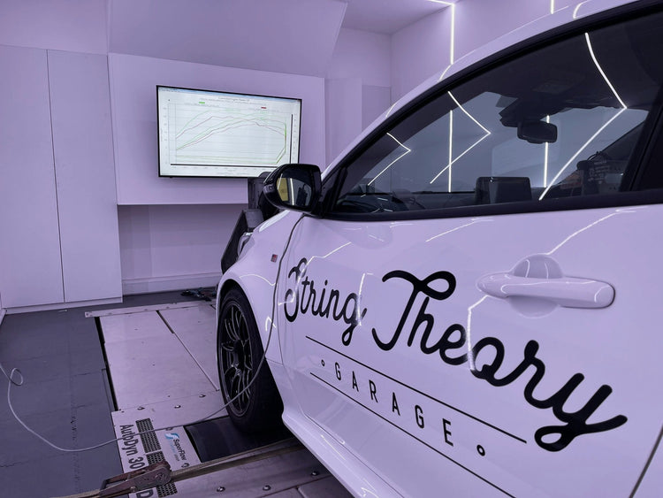 Side view of a white Toyota GR Yaris with the String Theory Garage sign writing on the passengers door, on a rolling road showing the data output on a large screen in front