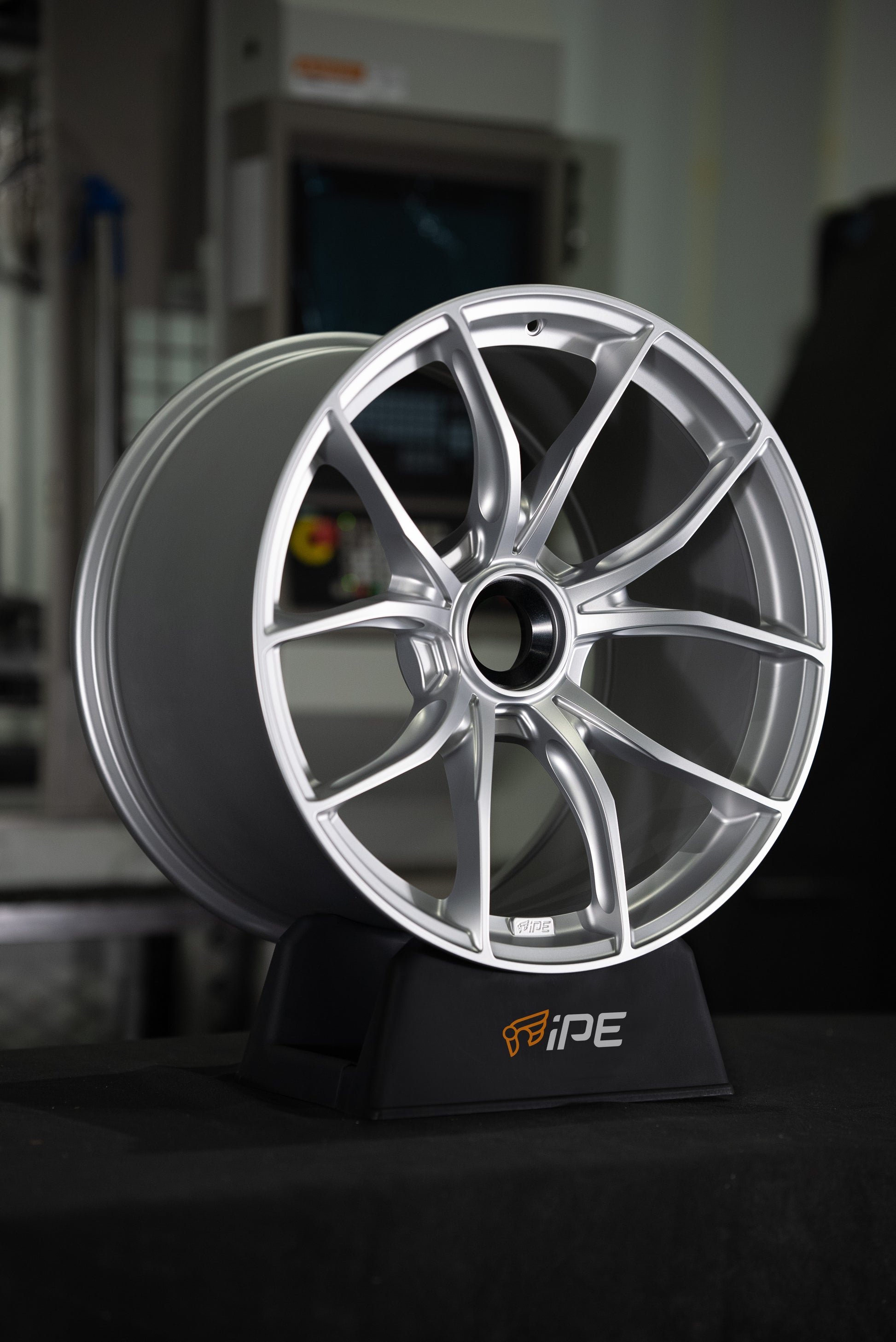 Front side view of an IPE MFR-01 Magnesium wheel in silver with a multi-spoke design sitting on an IPE stand with a workshop setting in the background