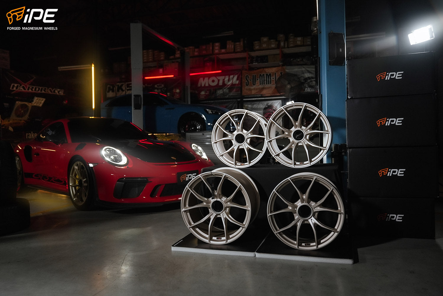 The photo depicts a dynamic automotive scene inside a well-lit garage. On the right, a stack of high-end, silver IPE wheel rims with intricate multi-spoke designs is showcased on a black display with the iPE logo. In the background, a vibrant red Porsche with black details sits prominently. The garage itself is lined with automotive parts and branding. The overall composition combines the allure of advanced auto parts with the prestige of sports car culture.