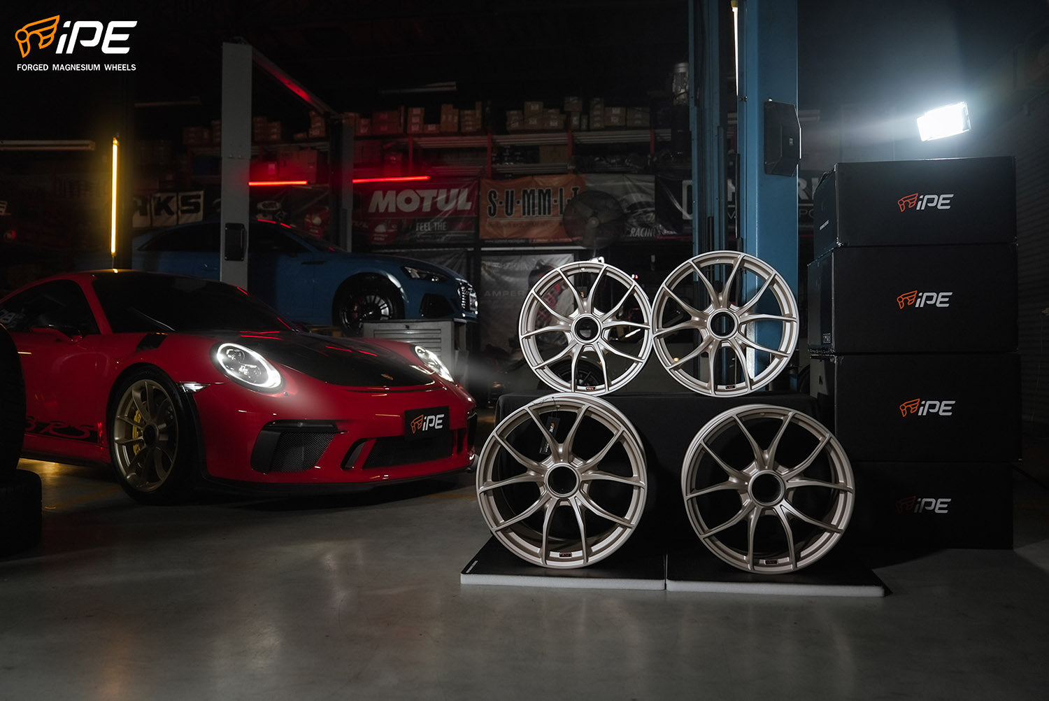 The photo depicts a dynamic automotive scene inside a well-lit garage. On the right, a stack of high-end, silver IPE wheel rims with intricate multi-spoke designs is showcased on a black display with the iPE logo. In the background, a vibrant red Porsche with black details sits prominently. The garage itself is lined with automotive parts and branding. The overall composition combines the allure of advanced auto parts with the prestige of sports car culture.
