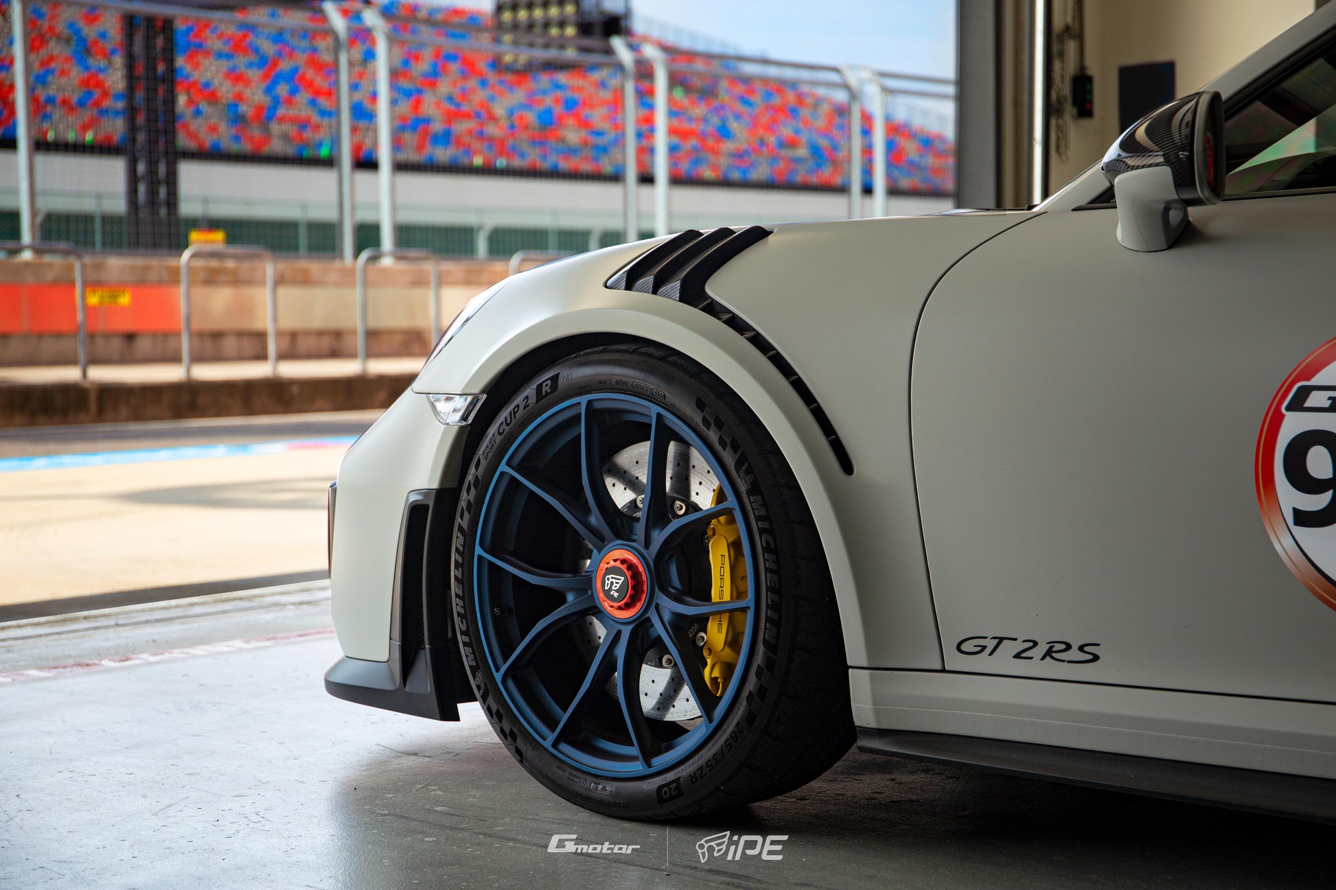 Nearside front view of a grey Porsche GT2 RS with a blue IPE MFR-01 Magnesium wheel fitted, inside the pit garage facing out to the pitlane at a race track