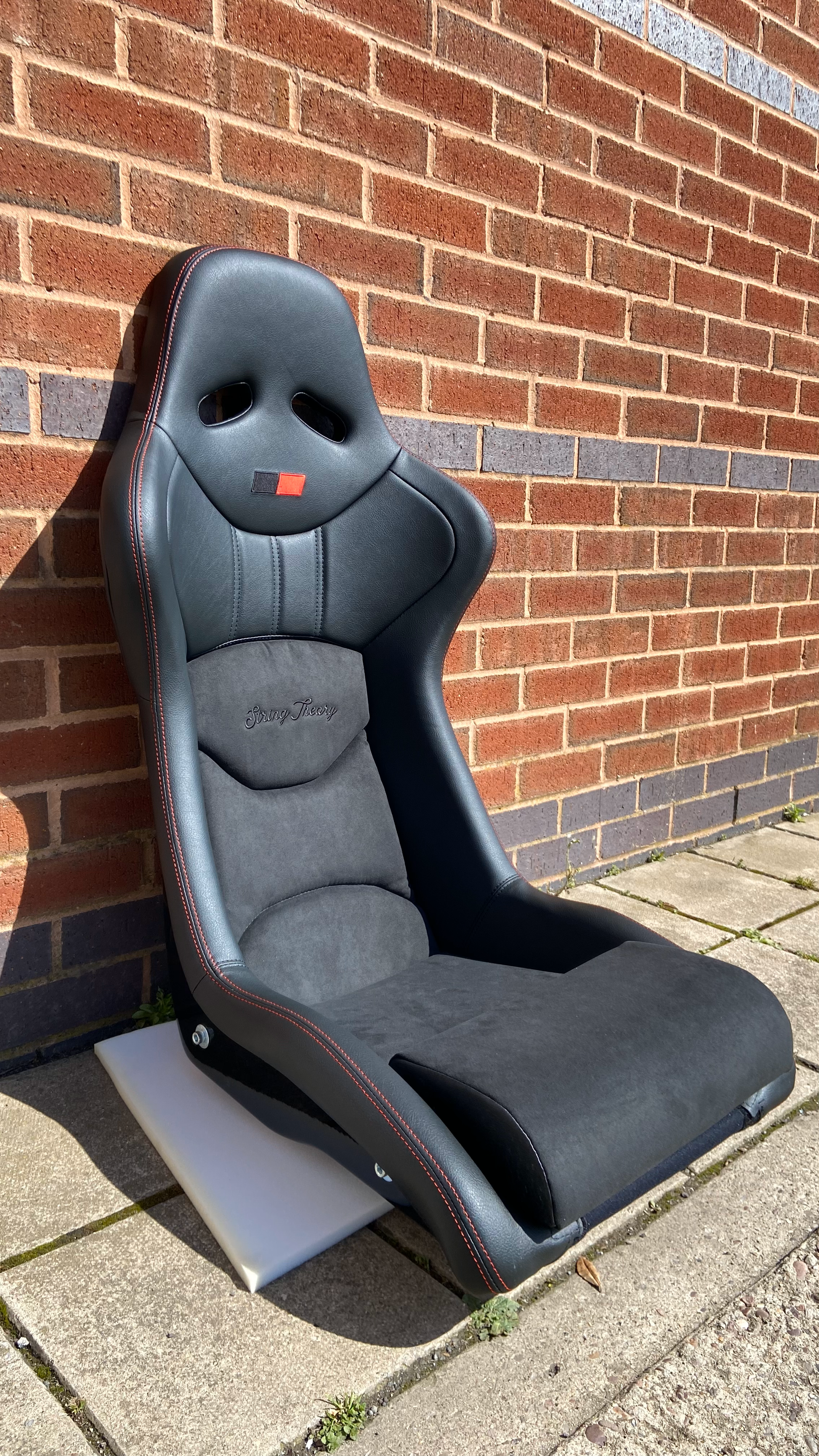 Offside front view of a black Cobra Nogaro Circuit sports seat with red stitching, red / black emblem on the headrest & a String Theory logo sewn into the back rest