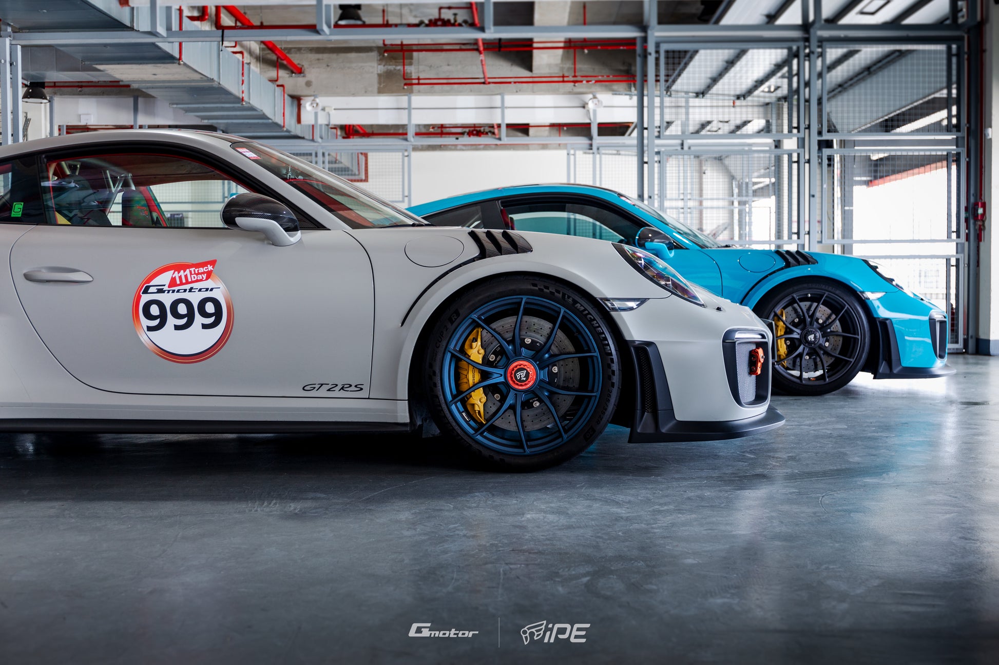 Side view of a grey Porsche GT2 RS with a blue IPE MFR-01 Magnesium wheel fitted side by side with a blue Porsche GT2 RS fitted with the same wheel, inside the pit garage facing out to the pitlane at a race track