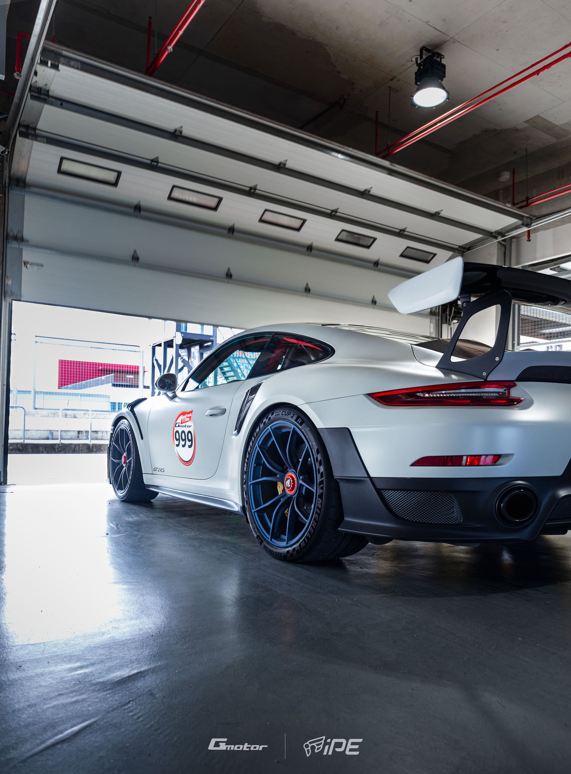 Nearside rear view of a grey Porsche GT2 RS with blue IPE MFR-01 Magnesium wheels fitted, inside the pit garage facing out to the pitlane at a race track