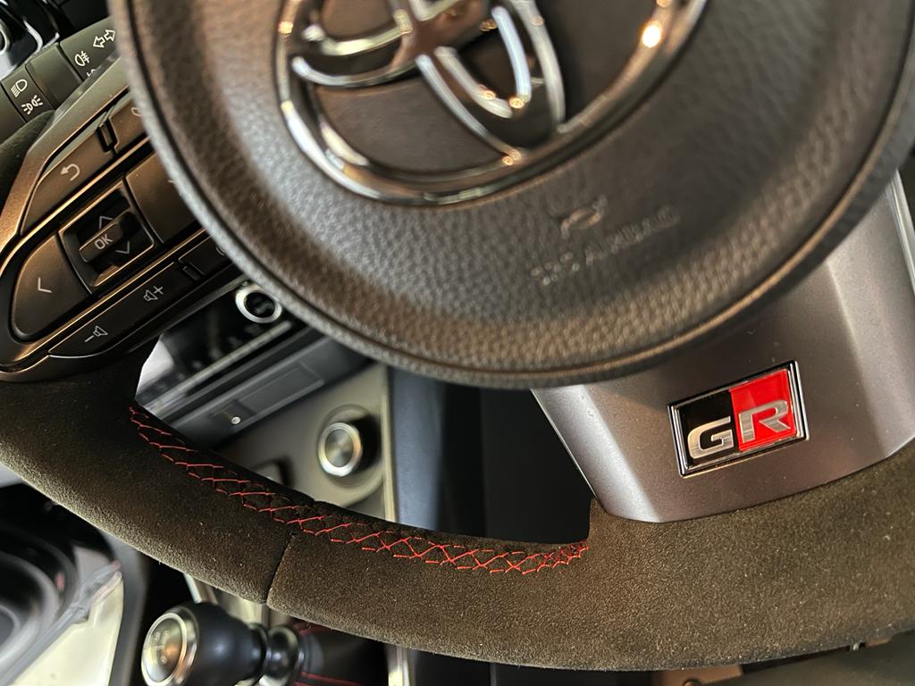 Extreme close up interior view of a Toyota GR Yaris fitted with a black Alcantara steering wheel with Red centre top stripe & stitching