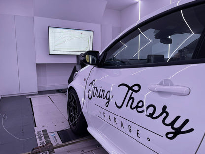 Side view of a white Toyota GR Yaris with black trim & String Theory Garage written on the door, on a rolling road showing performance figures on a screen in the background