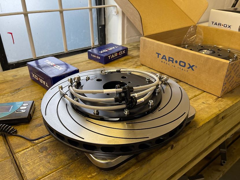 A Tarox vented & grooved brake disc with 3 braided brake lines on top, sitting on a set of weighing scales which is on a wooden workbench. There is another brake disc in a Tarox box to the right & two Tarox brake pad boxes in the background